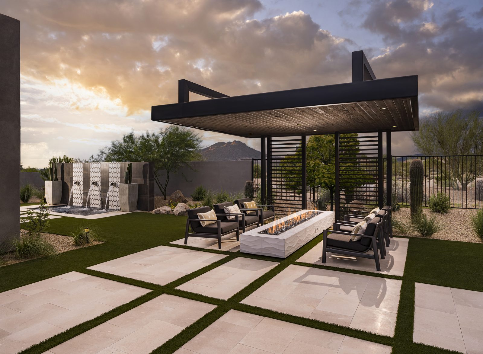 Travertine pavers with turf joints beneath a steel ramada and linear fire feature at dusk